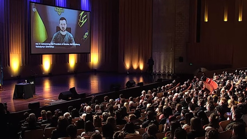 Zelenskyy's face is seen on a large screen overhanging a stage in a packed auditorium