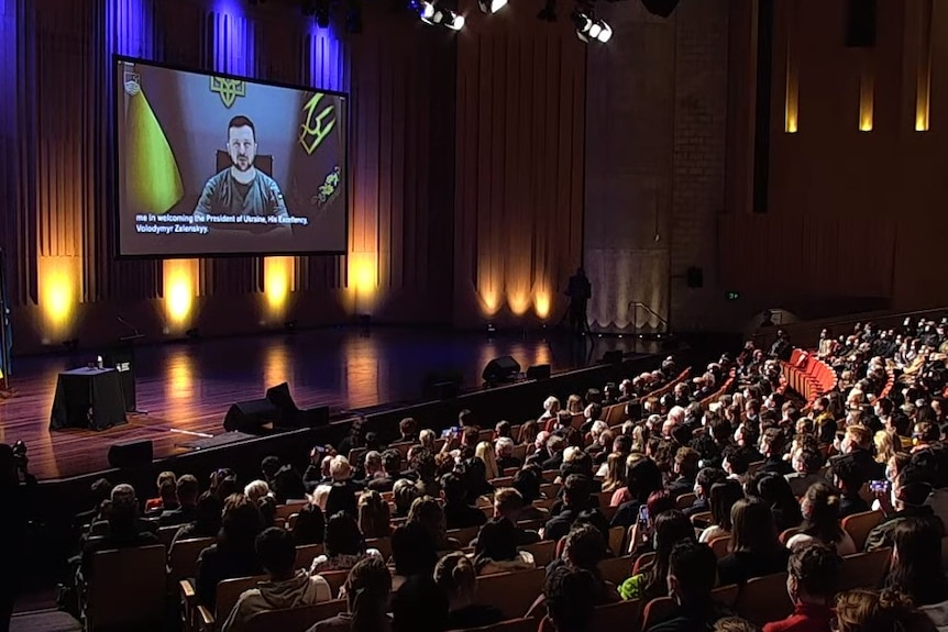 Zelenskyy's face is seen on a large screen overhanging a stage in a packed auditorium