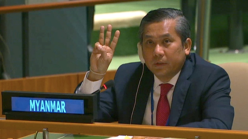 A middle-aged Asian man in dark suit, white suit & red tie, wears headset & holds up three fingers.