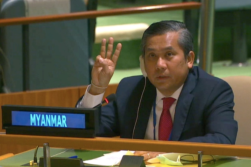 A middle-aged Asian man in dark suit, white suit & red tie, wears headset & holds up three fingers.