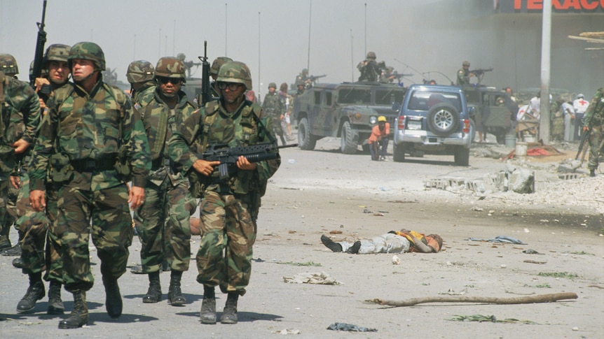 American troops walk past a Haitian man who lies dead in the street after a bomb was thrown into the crowd near a market.