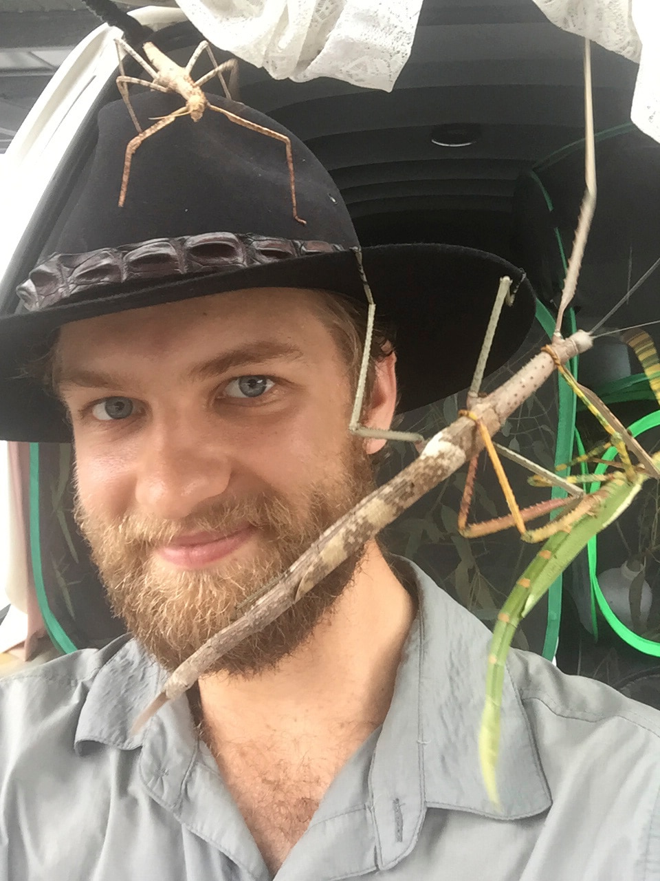 A smiling, bearded man in a dark, broad-brimmed hat holds a stick with a large insect clinging to it.