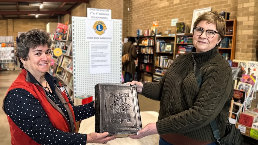 Sandie Morris wearing a red vest jacket presents an old bible to Nicole Penrose, wearing a dark green sweater in a bookshop
