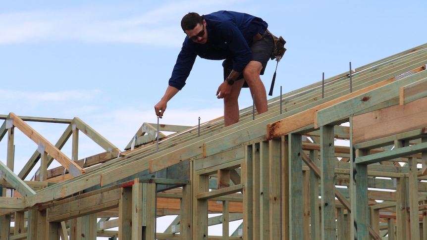 A builder (man) leaning over while standing on the top part of a house frame