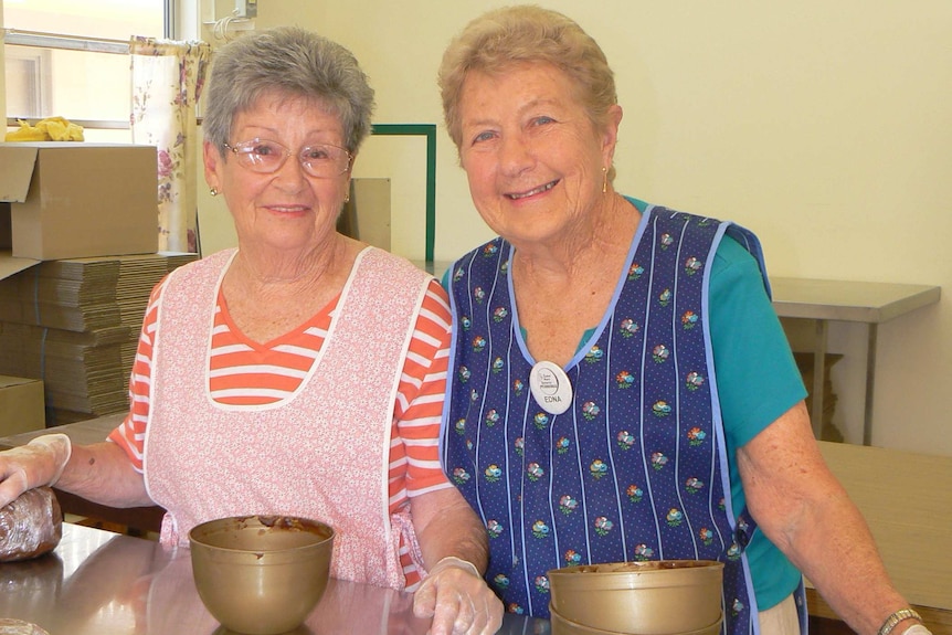 Two women wearing aprons stand in a kitchen with puddings in front of them wrapped in cling wrap