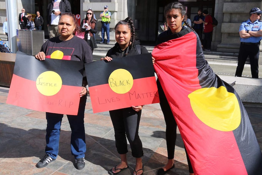 Three Aboriginal women stand holding Aboriginal flags with people and police officers in the background.