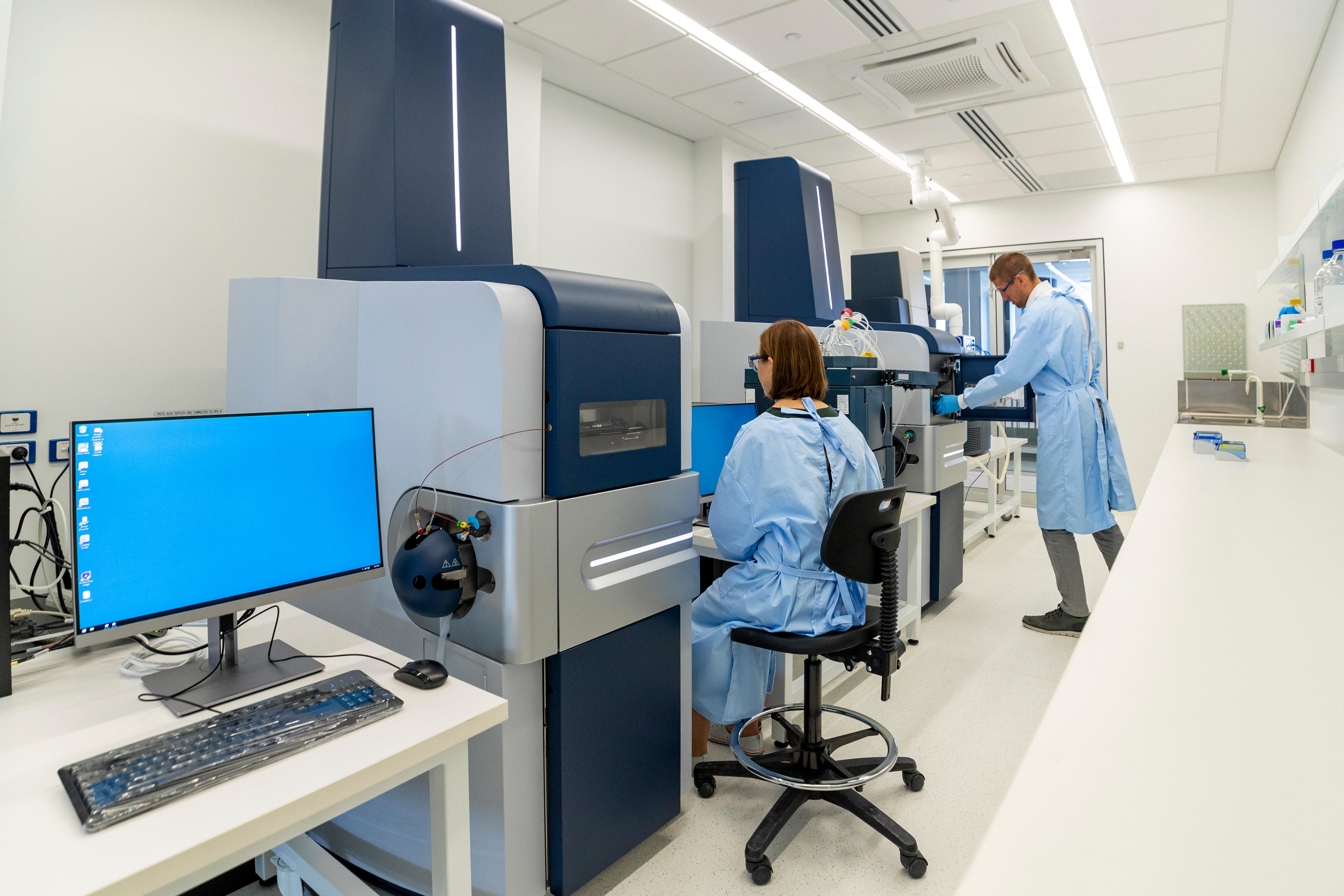 Two researchers in blue medical gowns conduct research inside room with large scientific machinery.