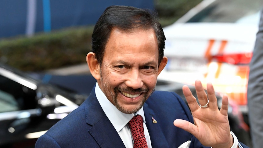 Sultan of Brunei Hassanal Bolkiah waves to a crowd, wearing a suit and tie.