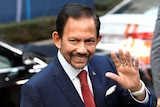 Sultan of Brunei Hassanal Bolkiah waves to a crowd, wearing a suit and tie.