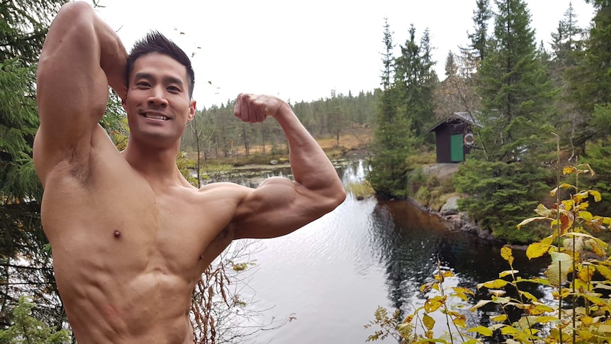 A muscular Viet Doan flexes his biceps next to a river surrounded by woodland trees.