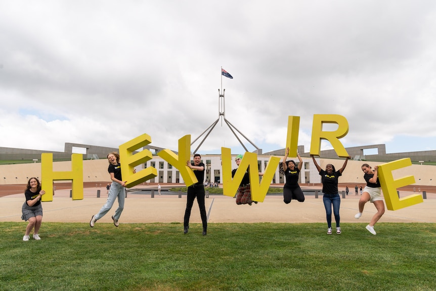 Seven young people holding big yellow letters that spell out 'Heywire' and jumping on grass in front of parliament house