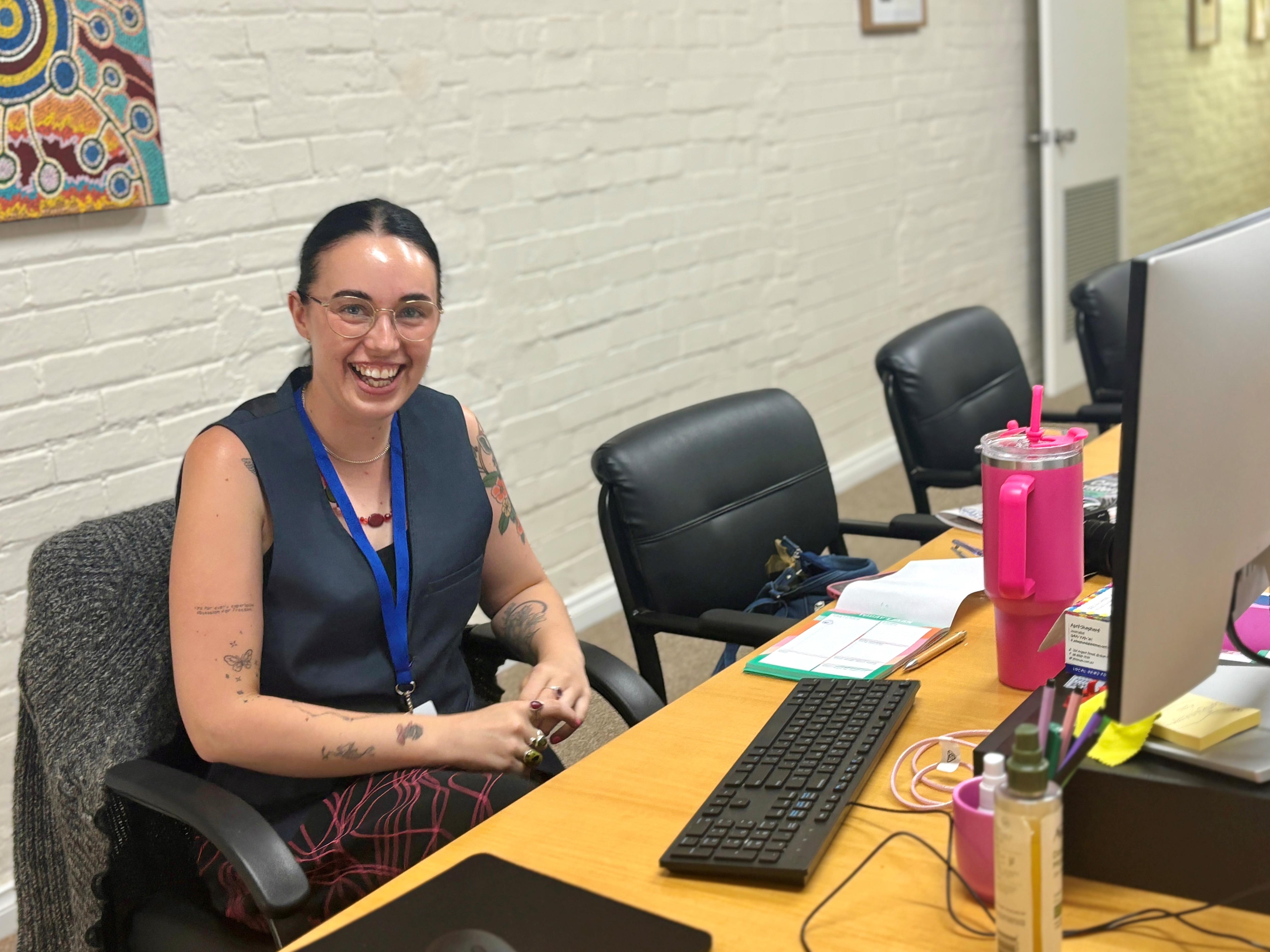 Una mujer con gafas sonríe desde un escritorio, tiene un vaso del tamaño de una jarra rosa, una computadora, viste una blusa azul sin mangas y un cordón alrededor del cuello.