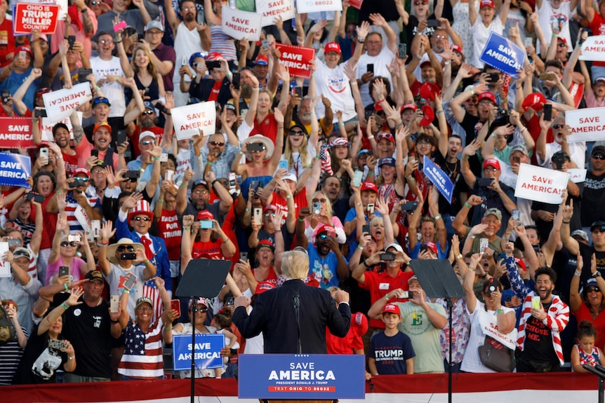 Donald Trump turns from his lectern to face a sea of cheering supporters on stadium seating behind him.