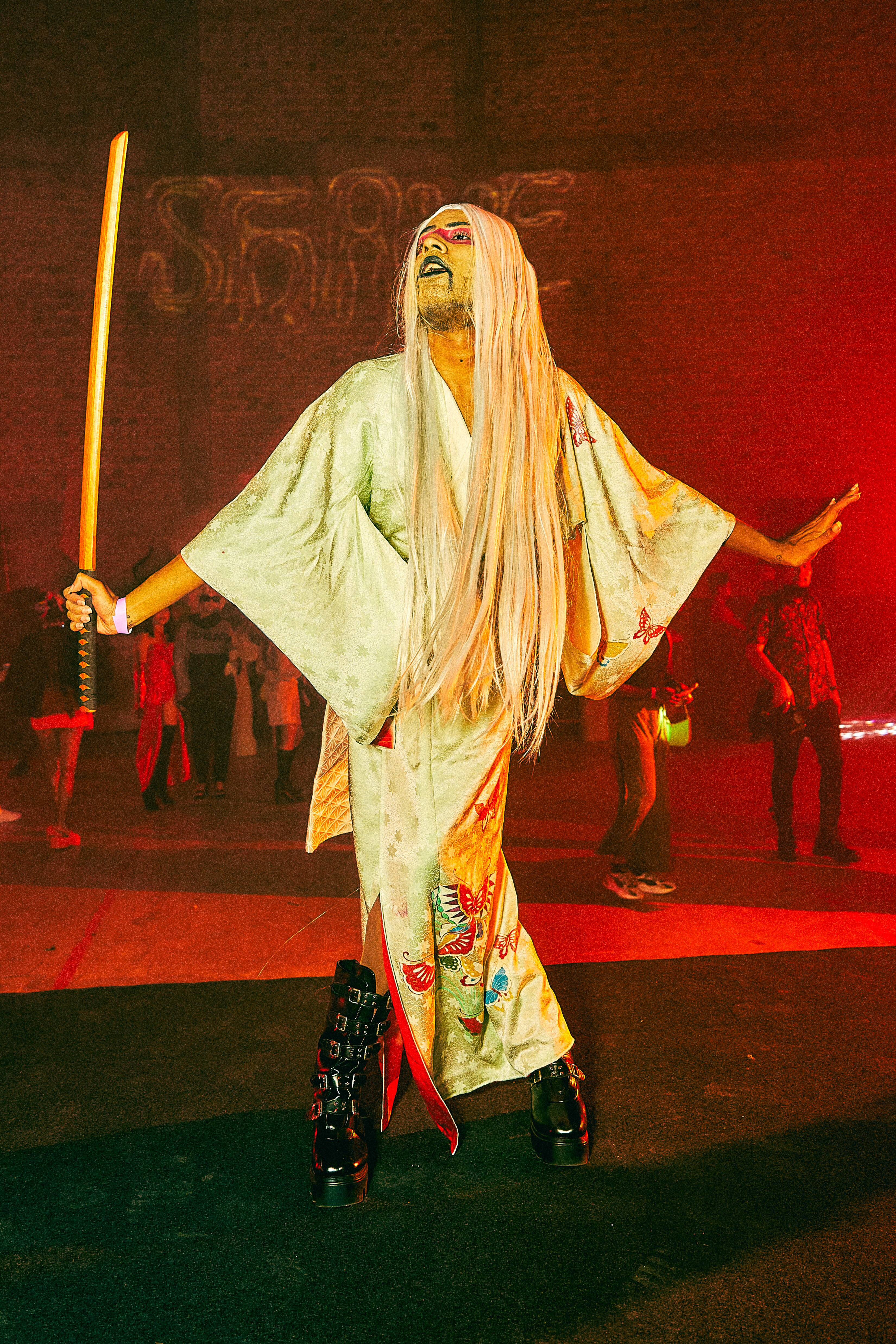 Miss BOOM posing in a long white wig, wearing a kimono-style outfit and holding a posing. 