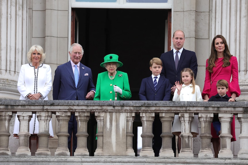 The royal family stands on a stone balcony. Charles and Camilla to the left of the Queen, William, Kate and brood to the right.