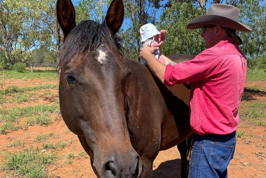 A man in a cowboy hat sits newborn baby, also in hat, on large horse.