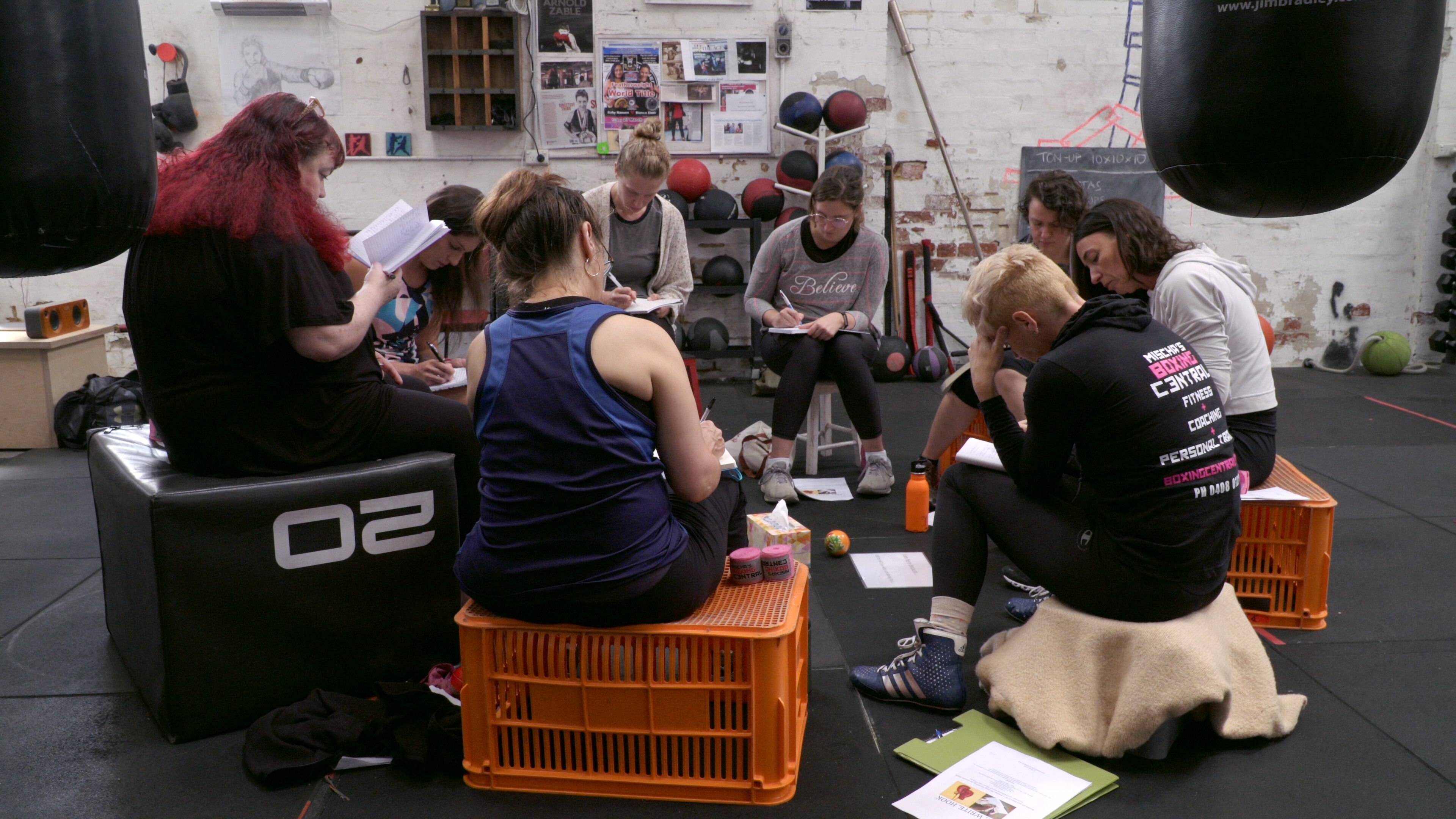 A group of women sit on crates and gym blocks, writing onto notepads. They are all heads down, deep in thought.