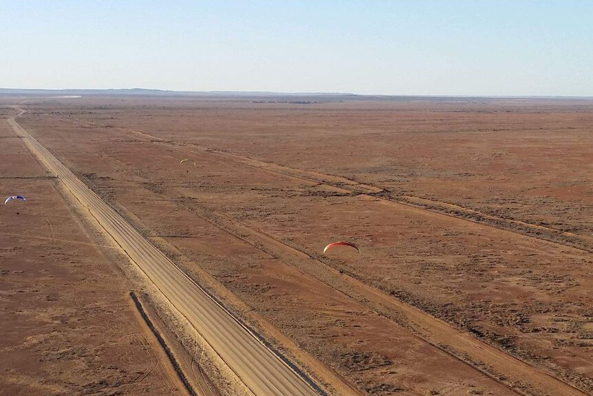 Three people can be seen paragliding across dry Australian terrain.