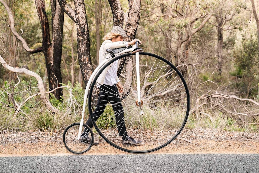 A man walking a penny-farthing bicycle along a road in bushland