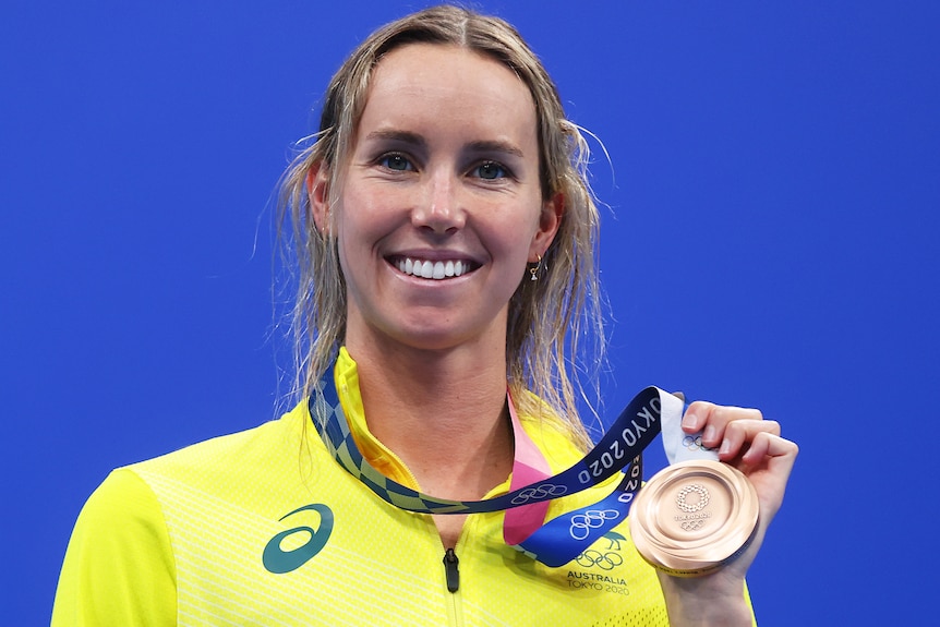 A blonde woman wearing a yellow jacket holding a bronze medal