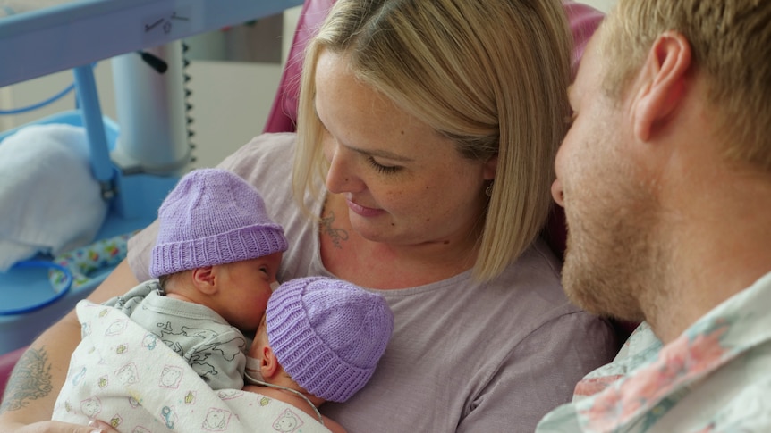 Tamara holding Tide and Kove on her chest, purple beanies on the babies, Justin looking over her shoulder.