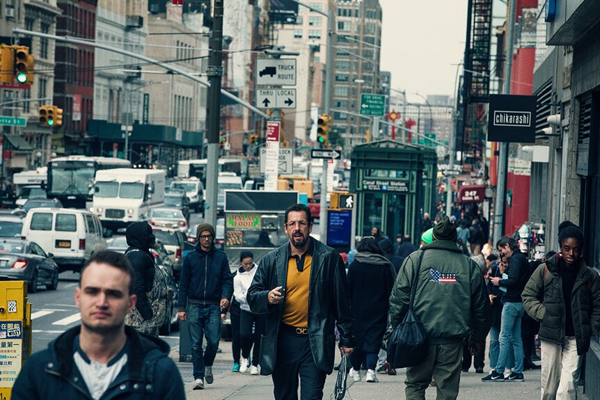 A man with black beard and short curly hair wears yellow polo, jeans, leather jacket and walks on busy New York City sidewalk.