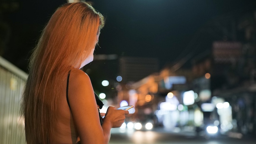 A woman with blonde hair looks at her phone on a street as shops and car lights shine behind her.