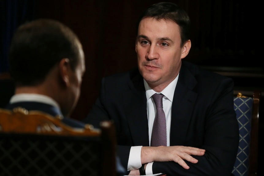 A man with dark hair wearing a black suit and patterned tie sits with his arms crossed