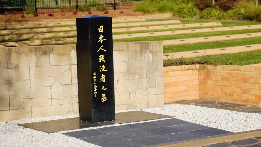 A black column with Japanese script stands in front of a low brick wall. Behind are rows with plaques on a lawn.