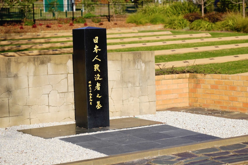 A black column with Japanese script stands in front of a low brick wall. Behind are rows with plaques on a lawn.