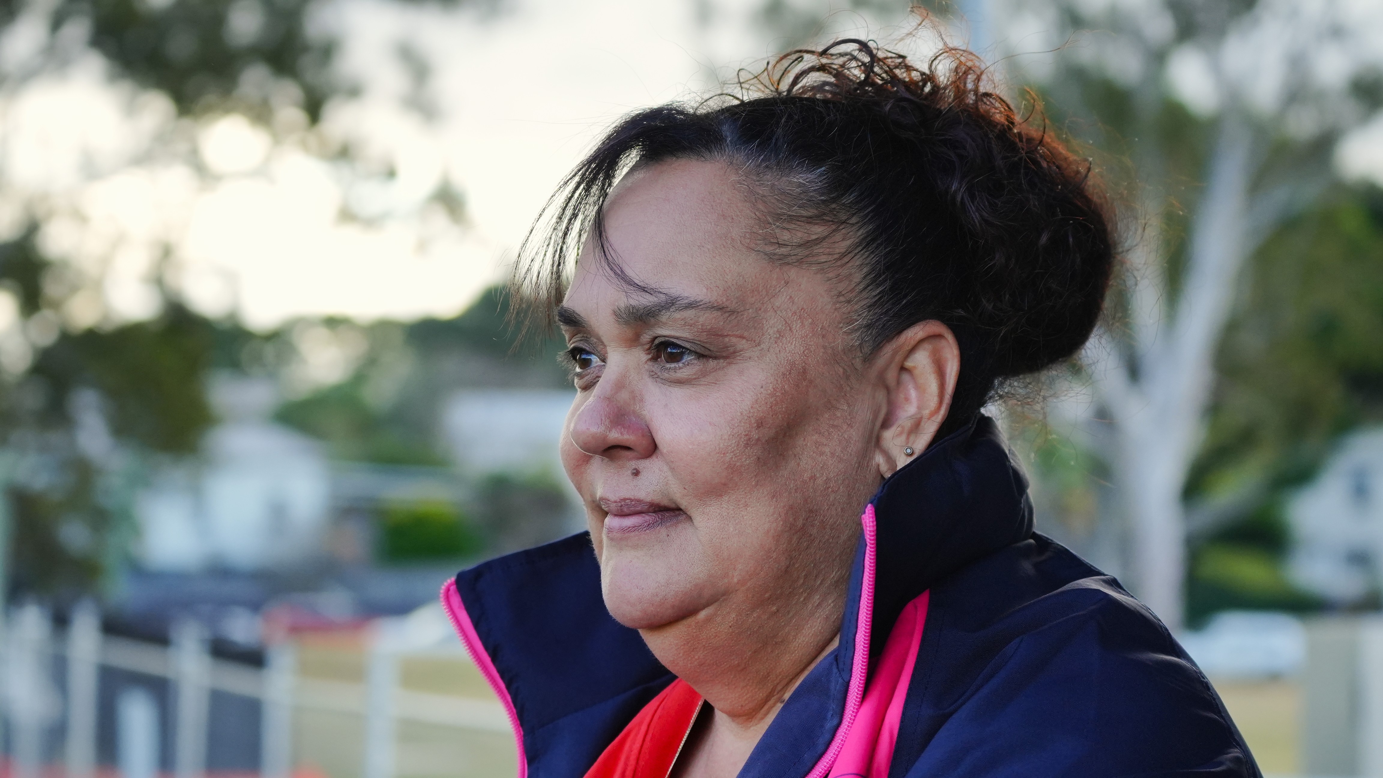 Mid-shot portrait of a tan-skinned woman dark hair looking off camera. There are gum trees behind her.