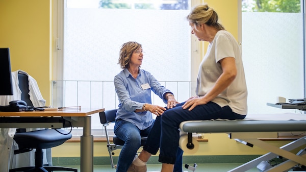 A woman with blue shirt and name tag sits looking up at and holding the knee of a woman who sits on a raised bed.