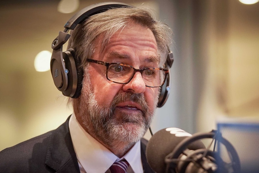 A man with a grey beard and glasses in front of a radio microphone.
