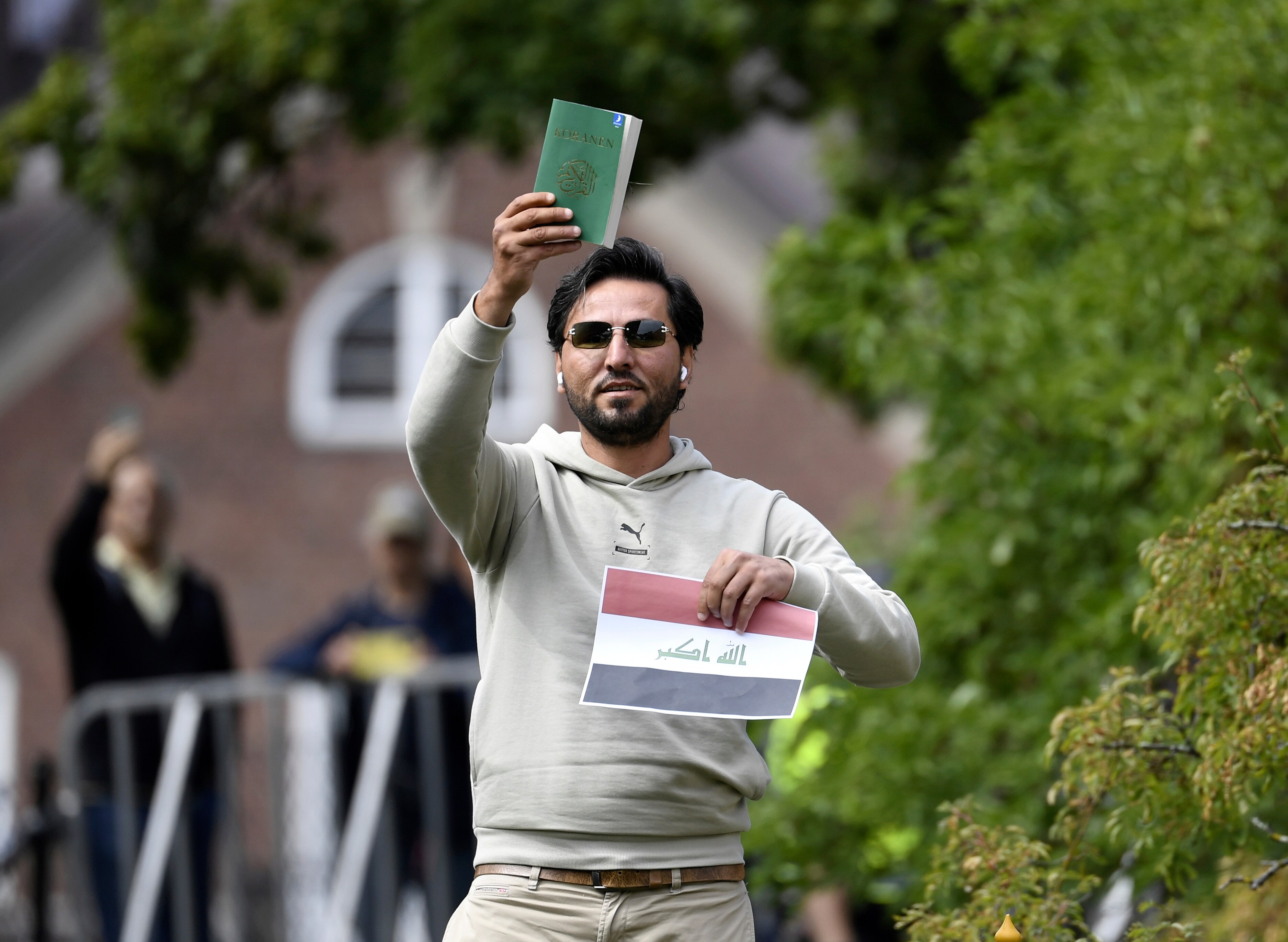 A man wearing sunglasses outside holding up a green book and a piece of paper with the iraqi flag printed on it.
