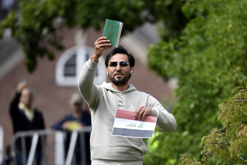 A man wearing sunglasses outside holding up a green book and a piece of paper with the iraqi flag printed on it.