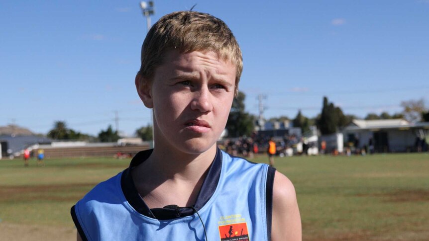 An adolescent boy with blonde hair and a blue football jersey looks past the camera.