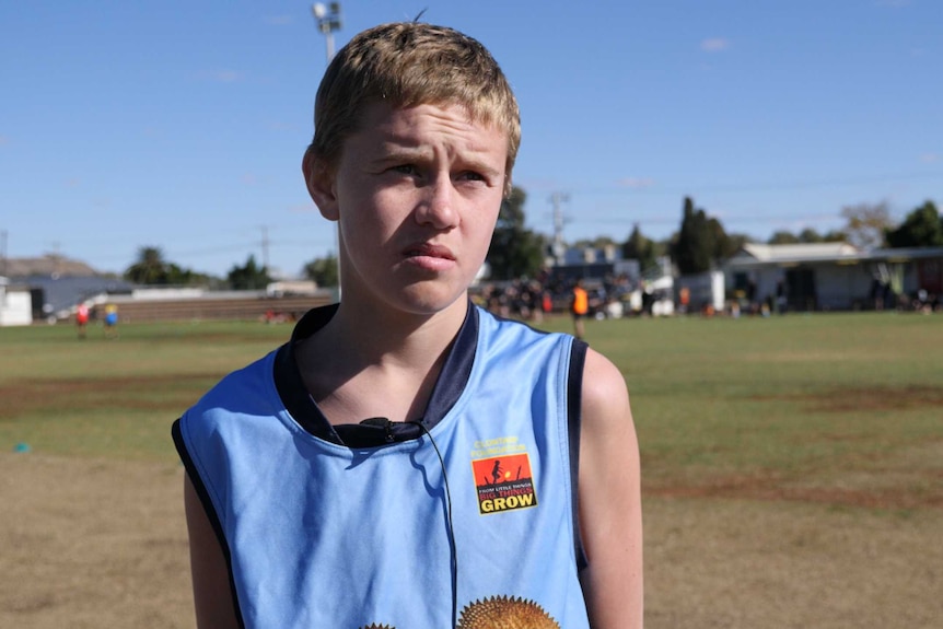 An adolescent boy with blonde hair and a blue football jersey looks past the camera.