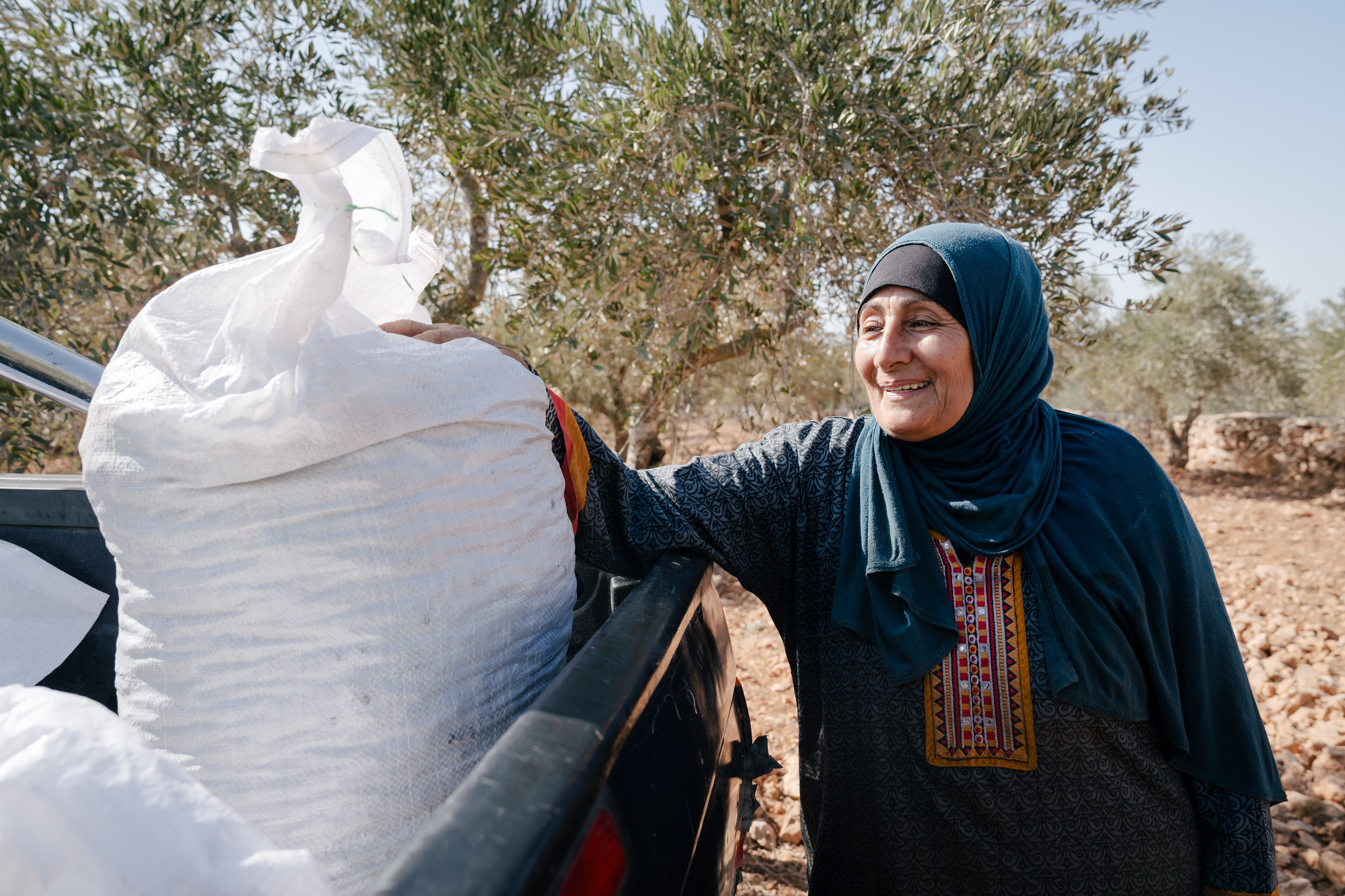 A woman wearing a hijab stands near the back of a utility truck with her hand on a big bag full of olives