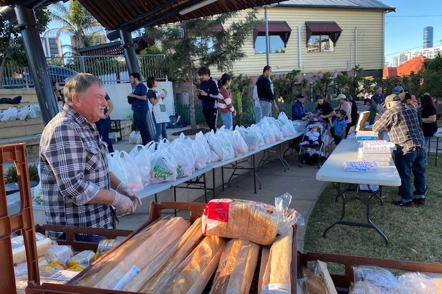 An elderly man stands ready to give out bread with a table full of bags of food behind him.