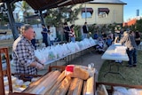 An elderly man stands ready to give out bread with a table full of bags of food behind him.