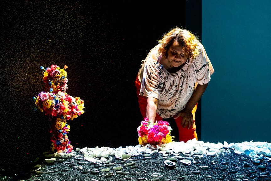 Elaine Crombie places a bunch of flowers on a mound covered in midden shells and a floral cross, in The 7 Stages of Grieivng
