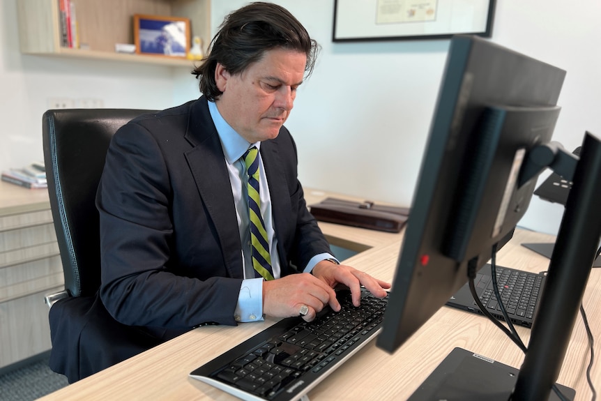 Innes Willox, wearing a dark suit, blue shirt and striped tie, sits at a desk and types on a computer. 