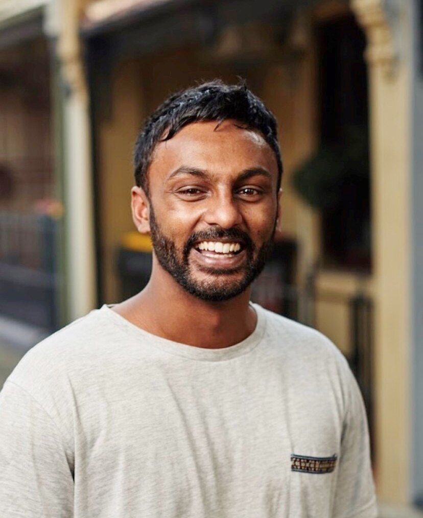 A portrait of a man in a white t-shirt smiling widely at the camera