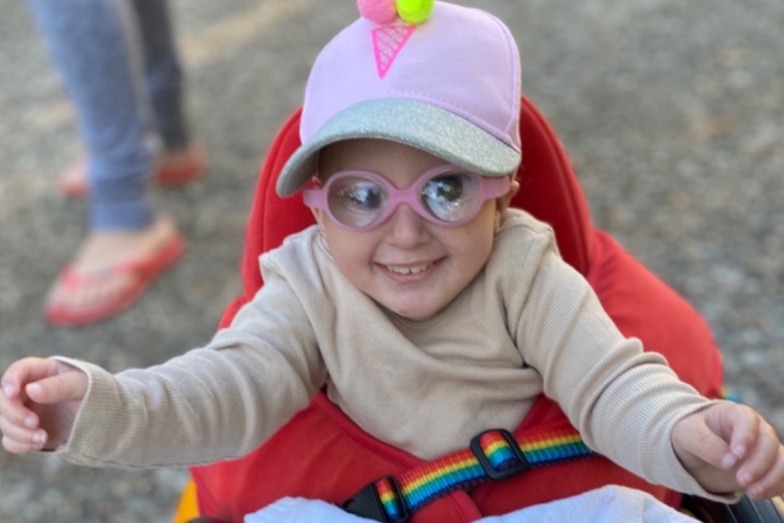 A tiny, smiling girl wearing a cap and pink glasses, holding her arms out.