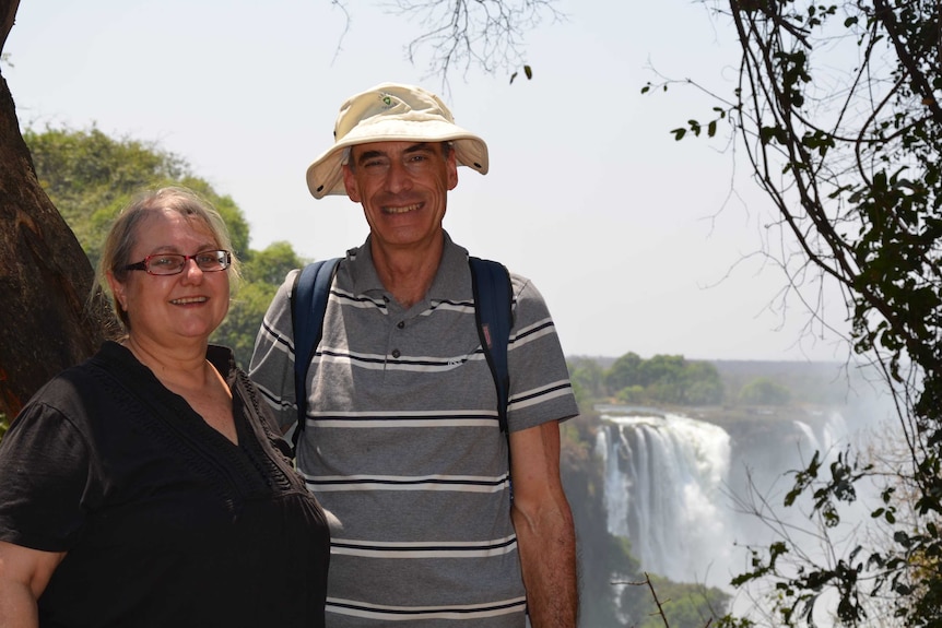 A woman with glasses and a man wearing a hat stand in front of a waterfall, both smiling