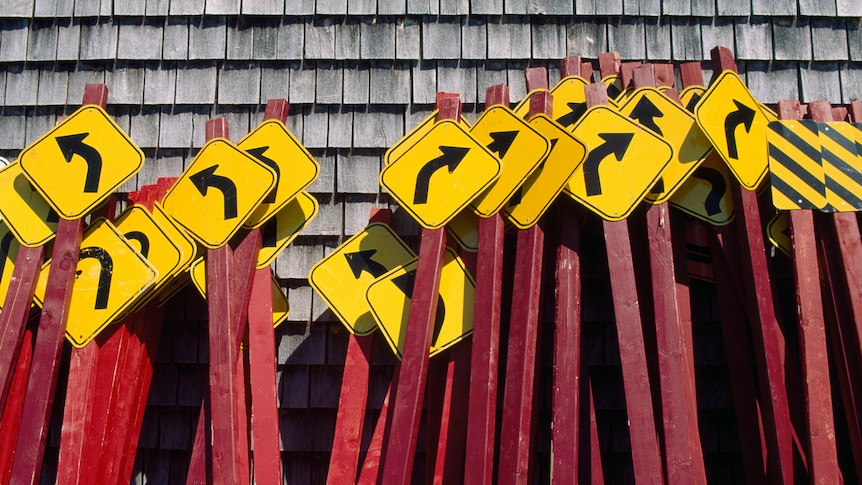 many traffic signs pointing in different directions leaning on a shed.