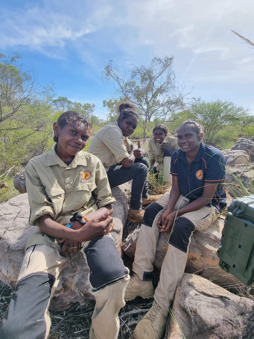 Four female rangers sitting on a rock in a bush environment, on a sunny day. They're smiling at the camera.