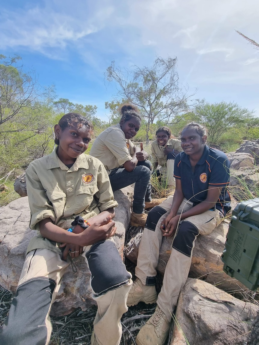 Four female rangers sitting on a rock in a bush environment, on a sunny day.  They're smiling at the camera.