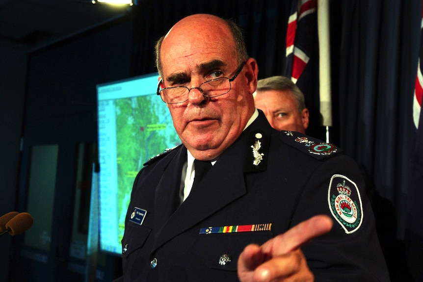 A man wearing a uniform and standing in front of an Australian flag points his finger.