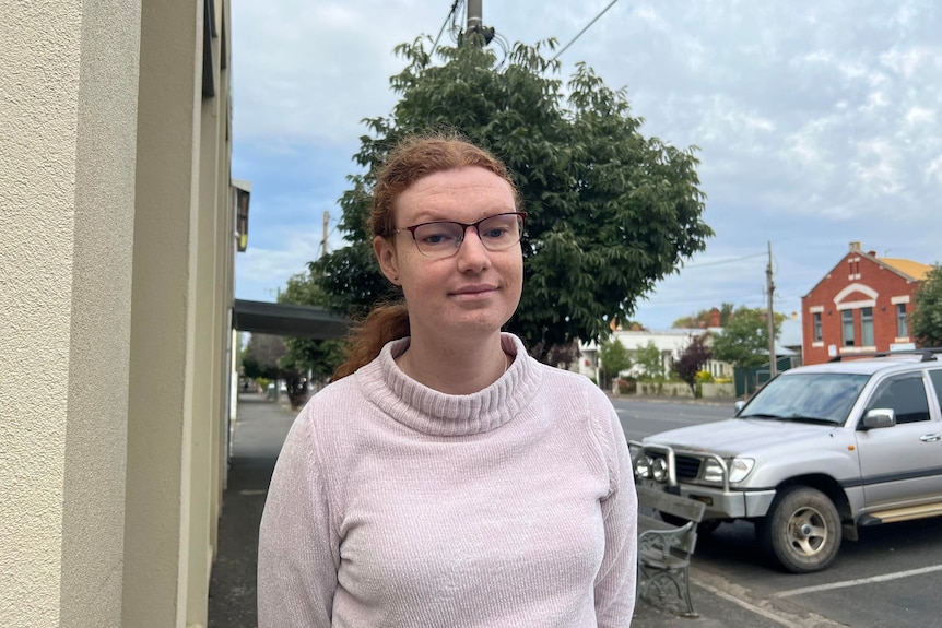 A ginger-haired woman with glasses stands on a street in a country town beneath an overcast sky.